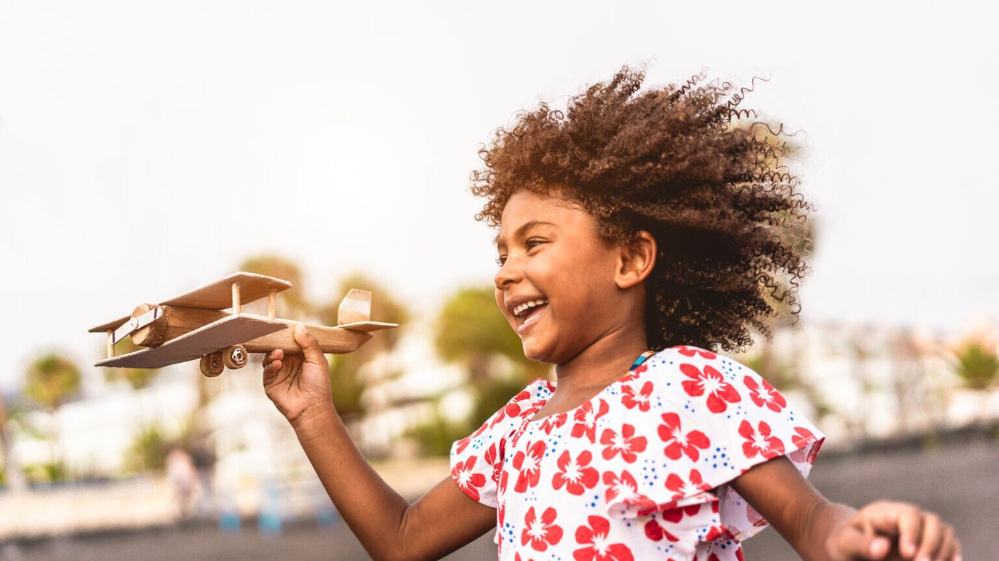 A young child running outdoors holding a wooden toy biplane above her head, laughing